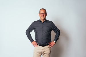 Roland Wulz has short hair, rectangular glasses, a dark grey shirt, beige trousers and has his hands on his hips.