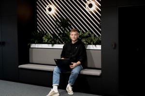Ein Mann mit schwarzem Kapuzenpullover und Laptop auf dem Schoß Hand sitzt auf einer gepolsterten Bank und lächelt in die Kamera. Hinter ihm befinden sich grüne Pflanzen.