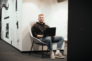 Ein Mann mit schwarzem Kapuzenpullover sitzt im Büro auf einem Stuhl mit einem Laptop auf dem Schoß