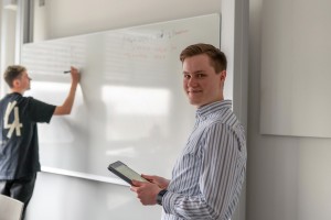 Valentin Müller doziert, schaut in die Kamera und ein Student steht an der Tafel und schreibt