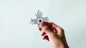 Eine Hand hält einen kleinen illustrierten Mikrochip mit Leitbahnen, die sich auf einem hellgrauen Hintergrund nach außen erstrecken